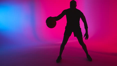 Studiosilhouette-Eines-Männlichen-Basketballspielers,-Der-Den-Ball-Vor-Rosa-Und-Blau-Beleuchtetem-Hintergrund-Dribbelt-Und-Hüpft